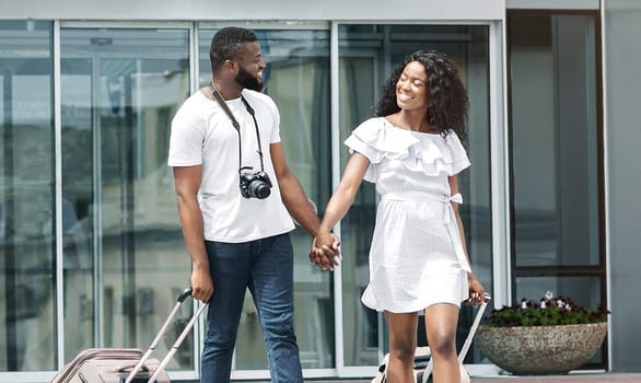 Black couple is seen walking hand-in-hand in front of a modern hotel building. They are both smiling and appear to be happy, possibly going on a vacation or business trip.