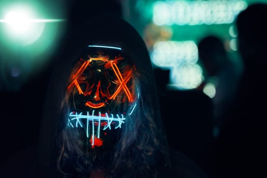 Human in hood with long disheveled hair in a lighting neon glowing mask. Anonymous man is a cyber criminal. Halloween dress-up party in night club. All Saints' Night.