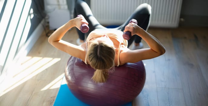 Top view of woman working out with dumbbells on fitness ball at home. Young female doing fitness exercise with dumbbells. Healthy lifestyle concept
