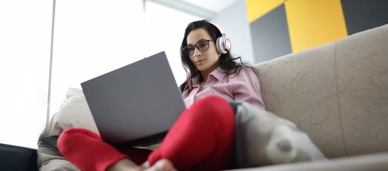 Portrait of woman sitting on sofa using laptop and headphones. Remote work or leisure time at home. Entertainment, distance learning or online meeting concept