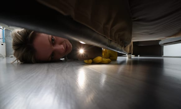 Close-up of woman using flashlight to look for something under bed. Female in protective gloves cleaning floor under bed