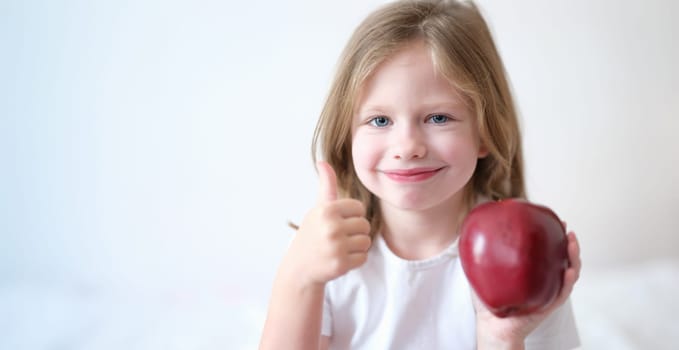 Portrait of little girl holding red apple in hands and showing thumbs up gesture. Healthy food and vitamins