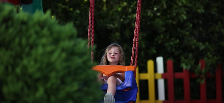 Portrait of little smiling girl riding swing on playground outdoors. Swing ride, cheerful child swinging. Happy kid having fun. Childhood concept