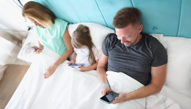 Top view of young family with kid using gadgets while lying together in bed. Leisure and fun time, happy family, parenthood and childhood