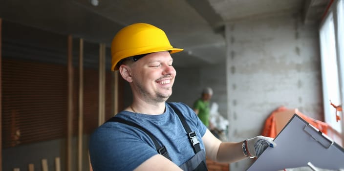 Portrait of smiling builder holding clipboard and writing down needed work material for renovation. Foreman in uniform and helmet. Building and construction concept