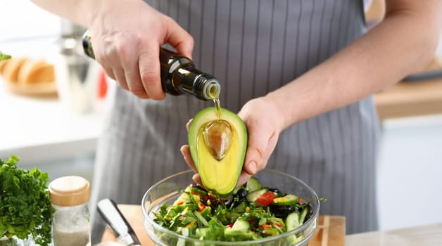 Close-up of cook holding bottle with olive oil, watering avocado with bone for cooking healthy salad. Nutritious meal raw food diet concept