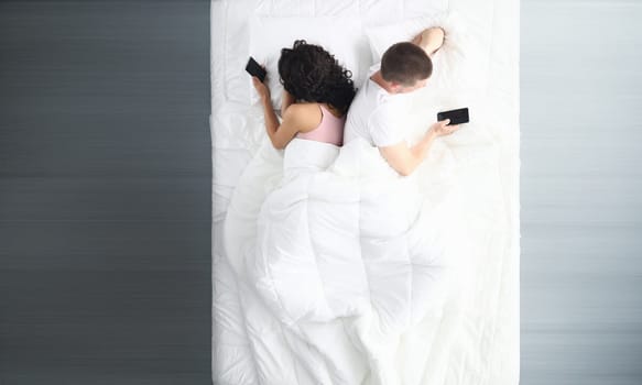 Top view of couple in bed using phones lying backs to each other. Man and woman surfing internet using cellphones. Relationship problems and virtual life