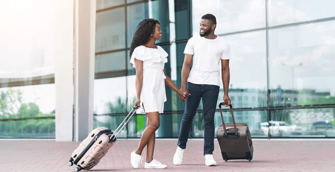 A smiling black couple, dressed in casual attire, walks through an airport terminal, holding hands and pulling their luggage behind them.