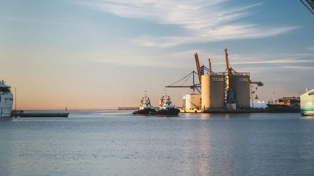 Sunset seaport scene with cargo ships, tugboats, cranes, and serene reflective water amidst industrial buildings. Calm yet functional maritime scenery download image