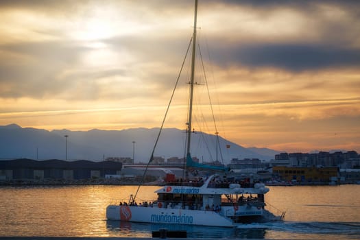 Spain Malaga 1 28 2024 Sailboat journey at sunrise sunset in 'Mundomarino'. Scenic view featuring shore buildings and distant mountains under golden light download image