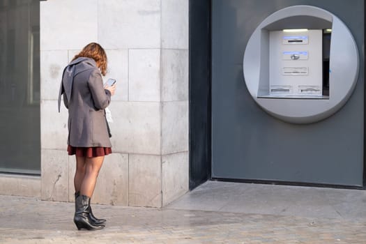 Urban dweller in stylish attire engrossed in smartphone near an outdoor ATM. Woman at Bank using smartphone. download image