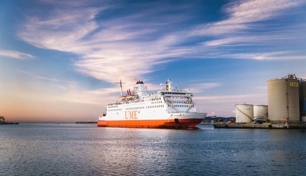 Large orange and white ferry boat anchored at port during sunset, with industrial structures visible in the background download image