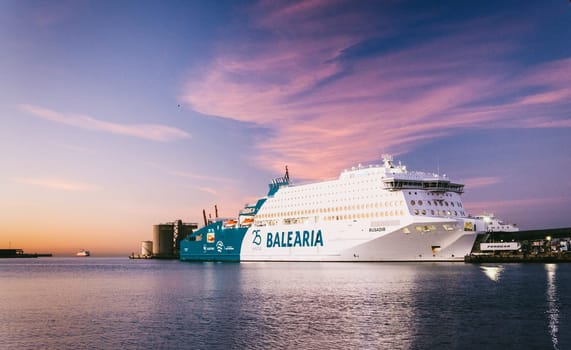 Sunset profile of BALEARIA ferry docked at bustling port, serene waters reflecting pinkish sky hues. Industrial background structures, second ship in view download image