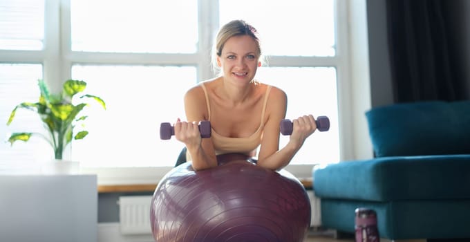 Portrait of smiling woman doing exercise with dumbbells on fitness ball. Fitness equipment for sport activity. Healthy lifestyle concept
