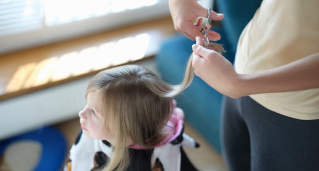 Top view of hairdresser making stylish hairstyle to cute little girl. Haircutter cuts and styles hair of small child. Fashion concept