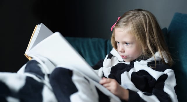 Portrait of little kid sitting on sofa and reading book. Distance education and home learning or leisure time at home