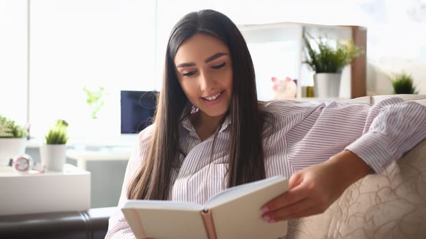 Portrait of young smiling woman reading book sitting on couch at home, interesting story. Cheerful brunette enjoy leisure time. Spare time and chilling concept