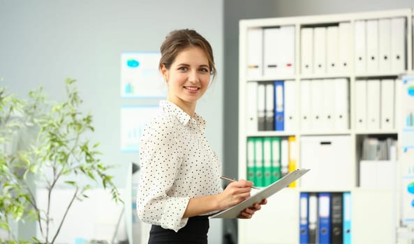 Portrait of cute smiling woman posing in conference room. Businesswoman holding clipboard with documents. Office interior on background