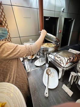 A woman at the buffet table in a hotel fills her plate with food from a food warming machine.
