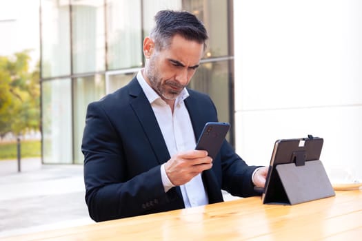 Satisfied with the results, the businessman sits outside the office building, a mature boss holds a phone and tablet writes messages and reads news online, using an app.