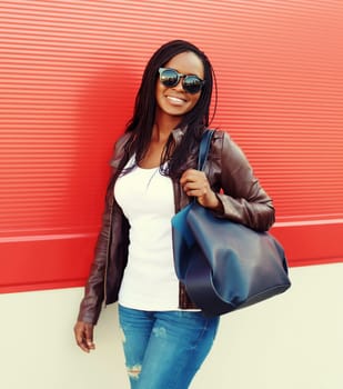 Fashionable happy young African woman with handbag in casual clothing, stylish modern black girl standing with dreadlocks, glasses posing in the city on red background