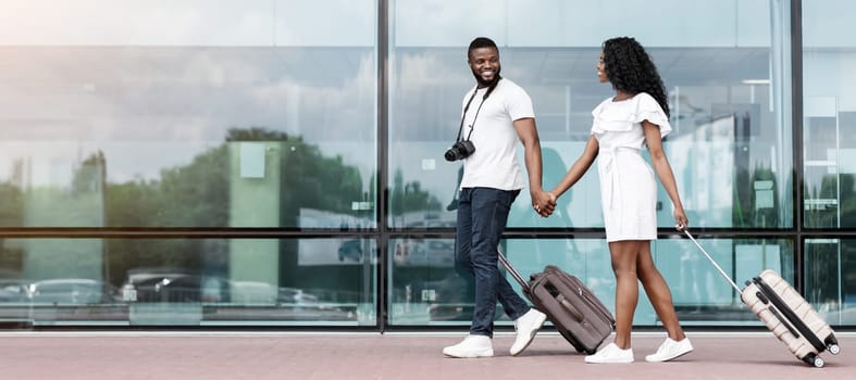 Black couple walks together through the airport terminal, holding hands and pulling their luggage behind them. They both appear to be smiling and happy, copy space