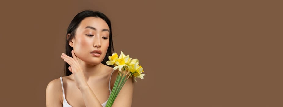 Young beautiful japanese lady posing with yellow daffodil flowers isolated on brown background, studio shot. Beauty and skin care, anti-aging treatment, spa procedure, ad and offer, copy space