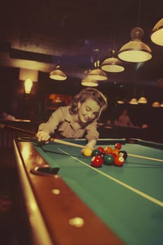 A woman is playing pool in a bar. She is wearing a white shirt and a blonde wig