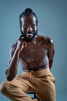 Smiling African American gay man with bright makeup. Shirtless muscular man sitting on stool and looking at camera. LGBTQ person is posing on blue background.