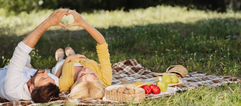Happy Couple Having Picnic Gesturing Heart Shape Lying On Plaid On Summer Date In Park Enjoying Day Together. Finally, Outdoor.