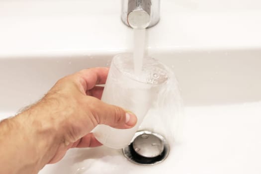 The left male hand holds a glass in the sink and water from the tap pours into the glass. Overflowing glass of water.
