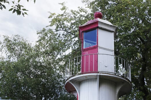 Top of coastal lighthouse krsn and white color, window, on the background of the sky and green foliage.