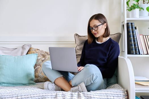 Young woman in glasses working at home, sitting on couch using laptop computer. Freelancing, training, education, remote work, leisure, internet online technology applications apps concept