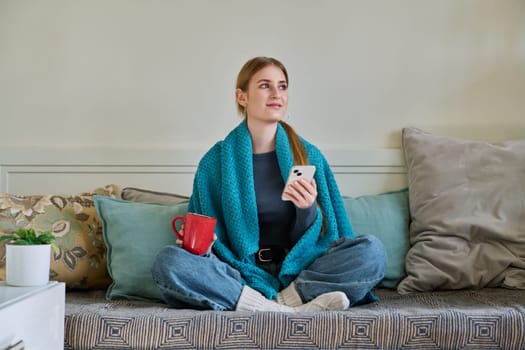 Young woman in warm clothes under blanket with cup of hot drink with smartphone sitting on couch at home, cold season, holiday weekend in winter. Lifestyle comfort coziness relaxation youth concept
