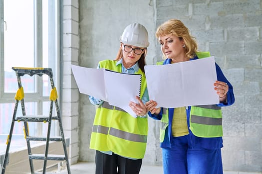 Two female industrial workers with technical plan at construction site. Serious female builders, engineers, inspectors, architects working with papers. Construction industry professions engineering
