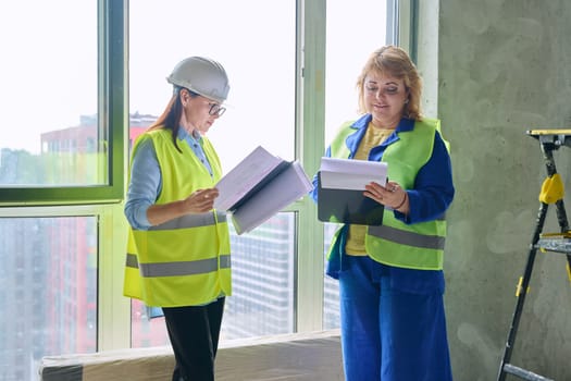 Two female industrial workers with technical plan at construction site. Serious female builders, engineers, inspectors, architects working with papers. Construction industry professions engineering