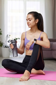 athletic fitness woman drinking water sitting on pink exercise mat at home