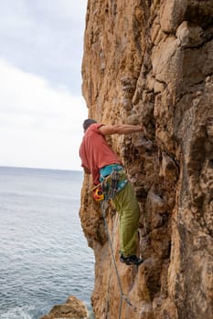 A man is climbing a rock wall with a rope around his waist. The scene is set against a backdrop of the ocean