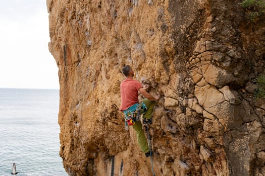 A man is climbing a rock wall with a rope. The man is wearing a red shirt and green pants