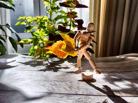 Small wooden figurine with a bouquet of autumn yellow birch leaves, bathed in sunlight, surrounded by shadows and backdrop of vegetation. Concept of autumn beauty and nature. Blurred, partial focus