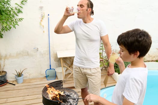 Father and son bonding during a summer barbecue by the pool. The father is drinking water while the son sips lemonade. Perfect family time and outdoor cooking concept.