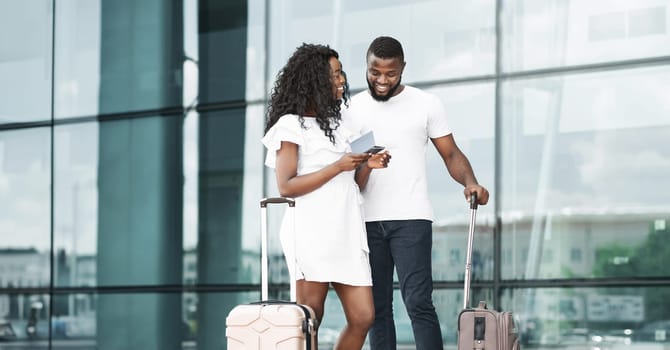 A happy black couple checking their boarding passes outside of an airport. They both have a luggage in their hands. They are smiling and looking at each other, seeming excited to start their vacation.