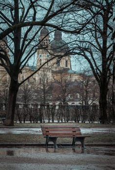 Munich, Germany - Dec 22, 2023 - Bench made wood and iron stand in the garden of the Munich Residenz with St.Cajetan and Adelaide in the background. Space for text, Selective focus.