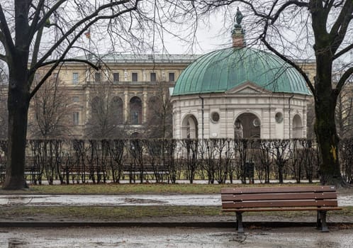 Munich, Germany - Dec 22, 2023 - Bench made wood and iron stand in the garden of the Munich Residenz with The Diana Temple (Dianatempel) and The Munich Residence Museum Building in the background. Space for text, Selective focus.