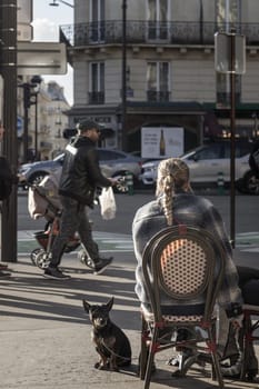 France, Paris - Jan 05, 2024 - A backshot of a Young Woman sits alone wooden chair with the her dog outside a cafe on street in Paris city. Lady drinking coffee and dog sitting near in an outdoor area of cafe/restaurant, Copy space, Selective focus.