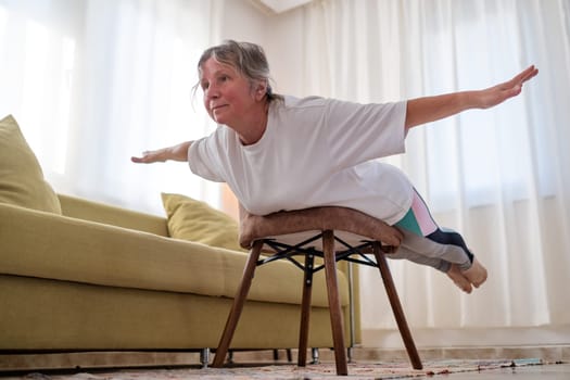 Senior caucasian woman doing yoga at home using chair as a props.
