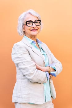 stylish mature entrepreneur woman smiling happy looking at camera leaning on an orange wall with arms crossed, concept of active lifestyle in adulthood