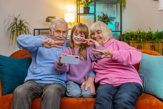 Happy grandfather, grandmother and granddaughter taking selfie photo with smartphone at home. Smiling Caucasian girl with grandparents make blogger self portrait showing peace sign sitting on sofa.