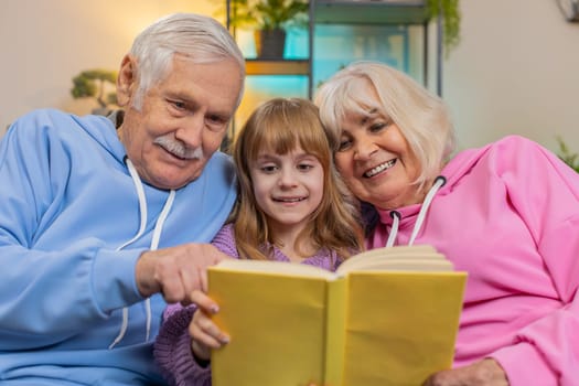 Caucasian grandparents and cute granddaughter reading interesting book together sitting on sofa in living room at home. Girl with grandparents spending leisure time on comfortable couch in apartment.