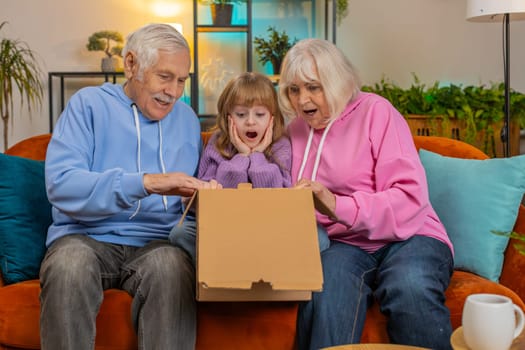 Happy amazed Caucasian grandparents and granddaughter girl opening cardboard box together sits on sofa at home. Smiling family consumers unpack good parcel looking inside great purchase delivered.